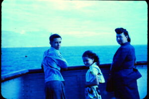 A young man, a girl, and their mother on a boat with calm waters in the distance.